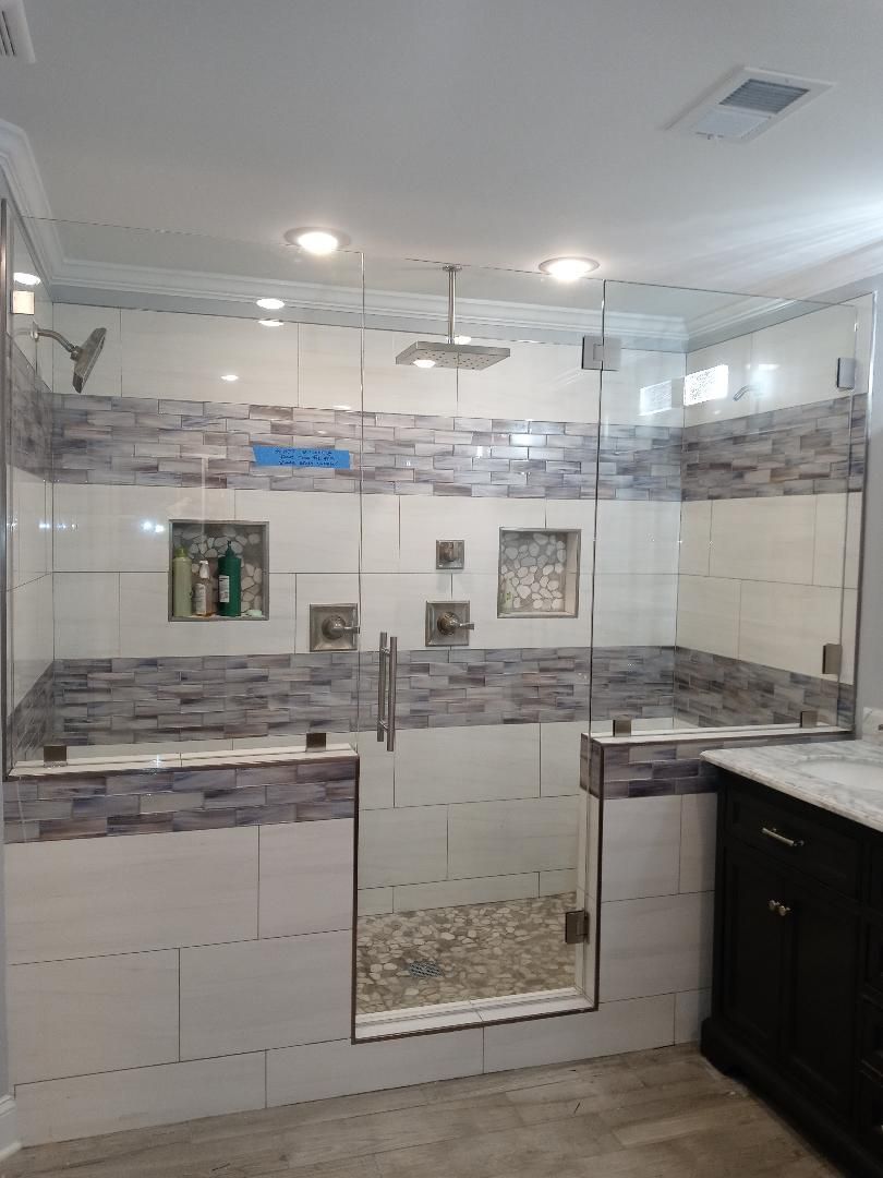 A newly renovated shower with glass doors and stone and white tile walls, including built-in shelving, a rain showerhead, and a pebble floor.