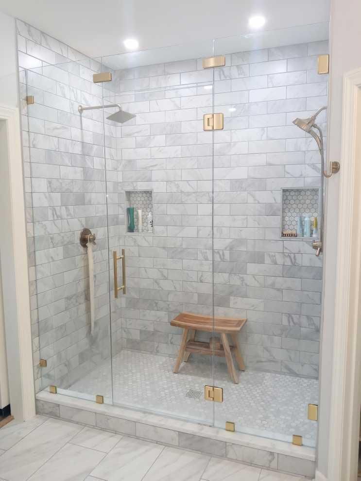 A luxurious walk-in shower with marble tile walls and floor, glass doors with gold hardware, and a wooden stool.