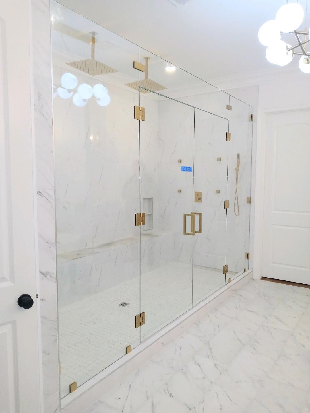 A luxurious shower with a glass enclosure, gold fixtures, and marble walls and floor. Bright white interior with natural light.