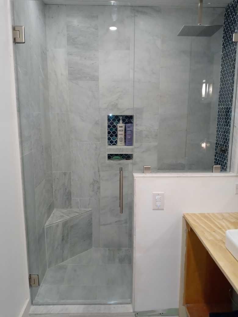 Glass-enclosed shower with gray marble-like tiles. Built-in niche with blue patterned tile holds toiletries. Includes a small corner seat.