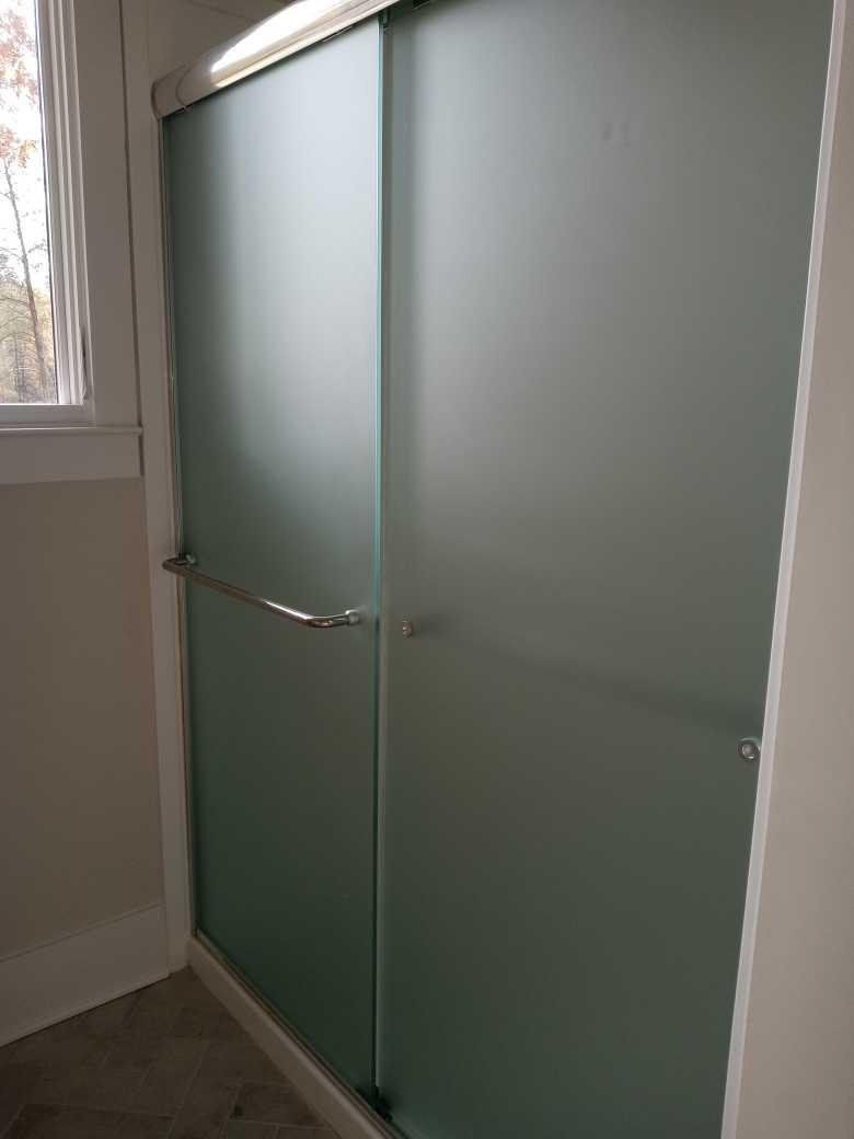 Frosted glass shower door with a silver handle, set against a white frame and wall, next to a small window.