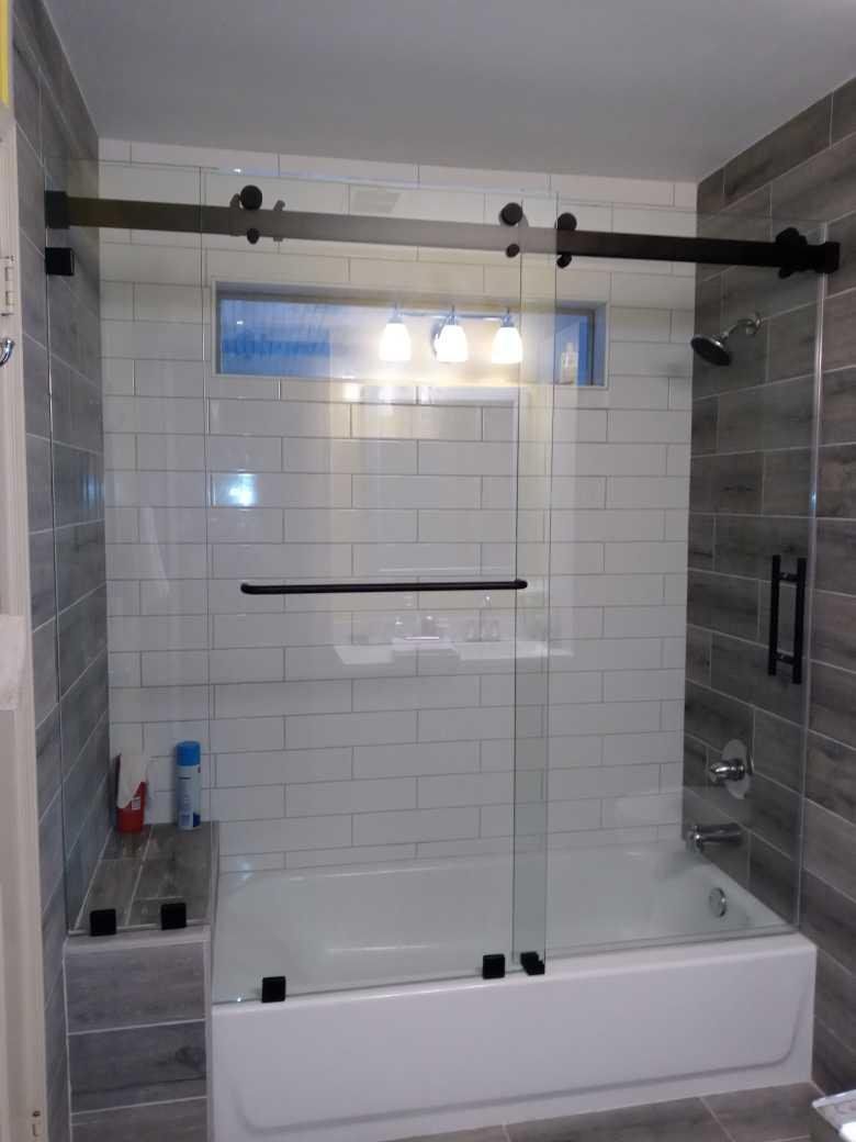 A modern bathroom with a sliding glass shower door over a white bathtub. Black hardware contrasts with the clear glass and light-colored tile.