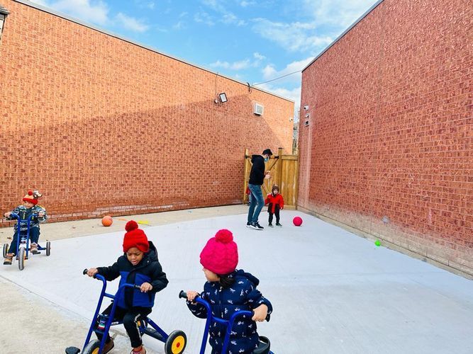 Children riding tricycles in a brick-walled courtyard. A man watches them, two balls lie on the ground.