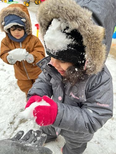 Two children in winter coats playing with snow. One with snow on their hood.