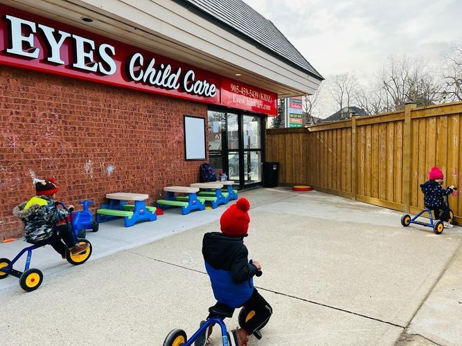 Children on trikes at Eyes Child Care, red building, wooden fence, outdoor play area.