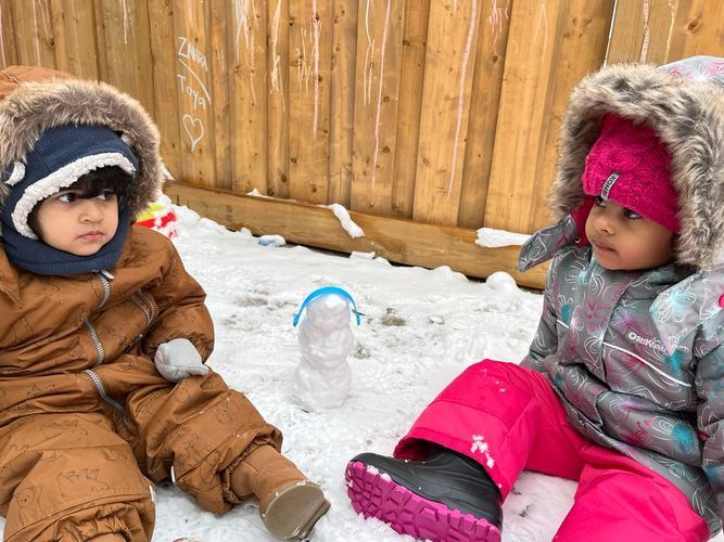 Two children in winter gear sitting in the snow near a small snowman, wooden fence in the background.