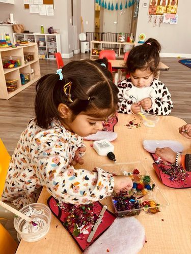 Two young girls at a table in a classroom, working on a craft with small colorful objects.