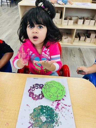 Girl in pink, surprised, at art table with stamped designs in classroom.