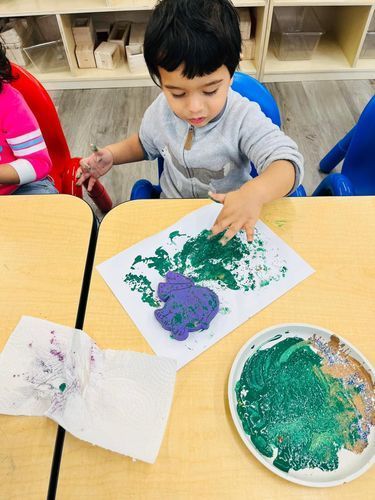 A young boy paints with his hands, using green and purple paint, at a table in a classroom.