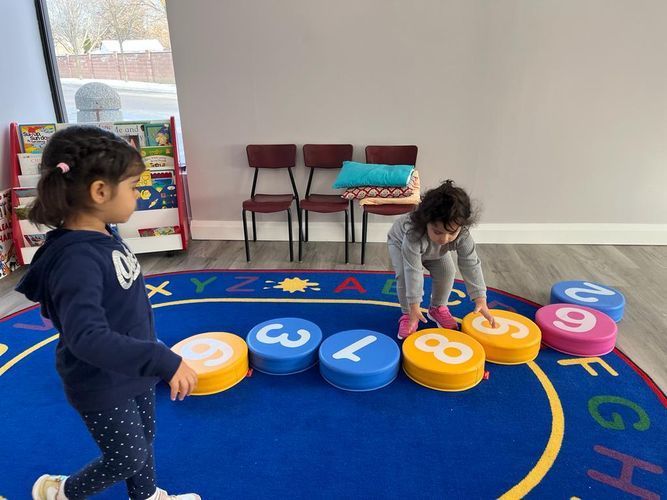 Two young girls playing with numbered floor cushions in a classroom setting.