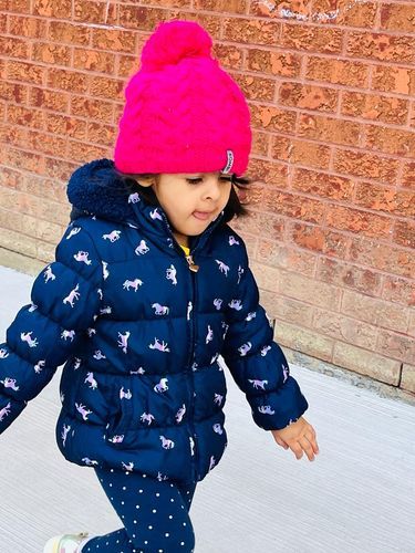 A young child in a pink hat and navy horse-print coat walks in front of a brick wall.