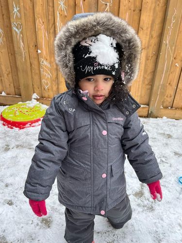 Child in gray winter coat and hat, with snow on their head, outdoors.