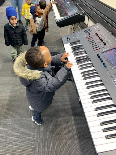 Young child playing a large keyboard, other children watch. Gray floor, dark coat, and keyboard.