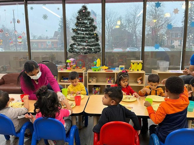 Children eating at a table with a teacher wearing a mask in a classroom with a Christmas tree.