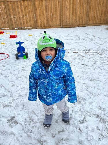 Young child in blue winter coat and dinosaur hat standing in snow.