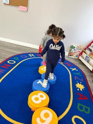 Two young girls stepping on numbered stepping stones on a blue alphabet rug in a classroom.