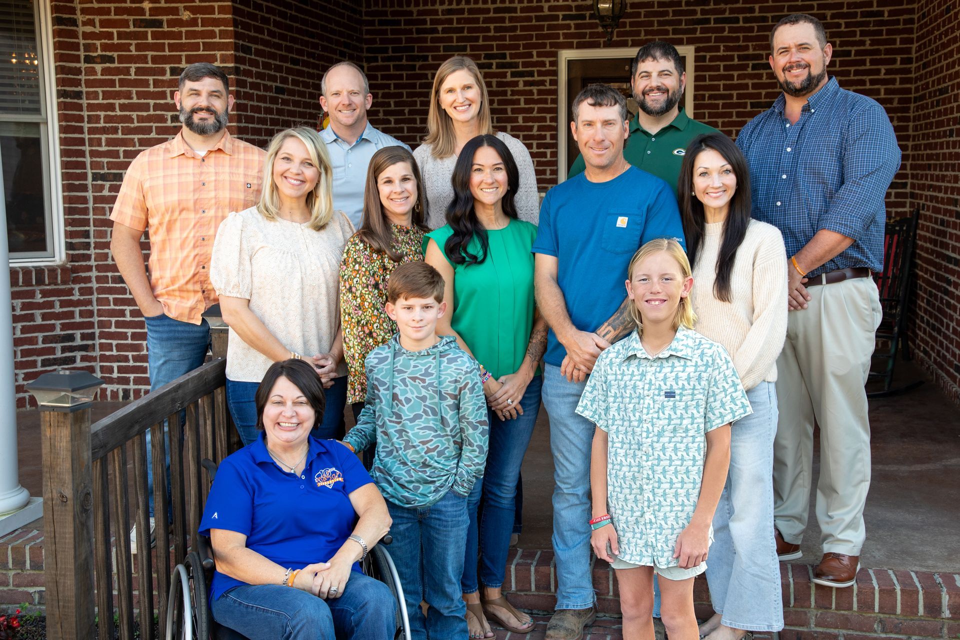 Group portrait on porch: people of various ages, brick background. Woman in wheelchair smiles at front.