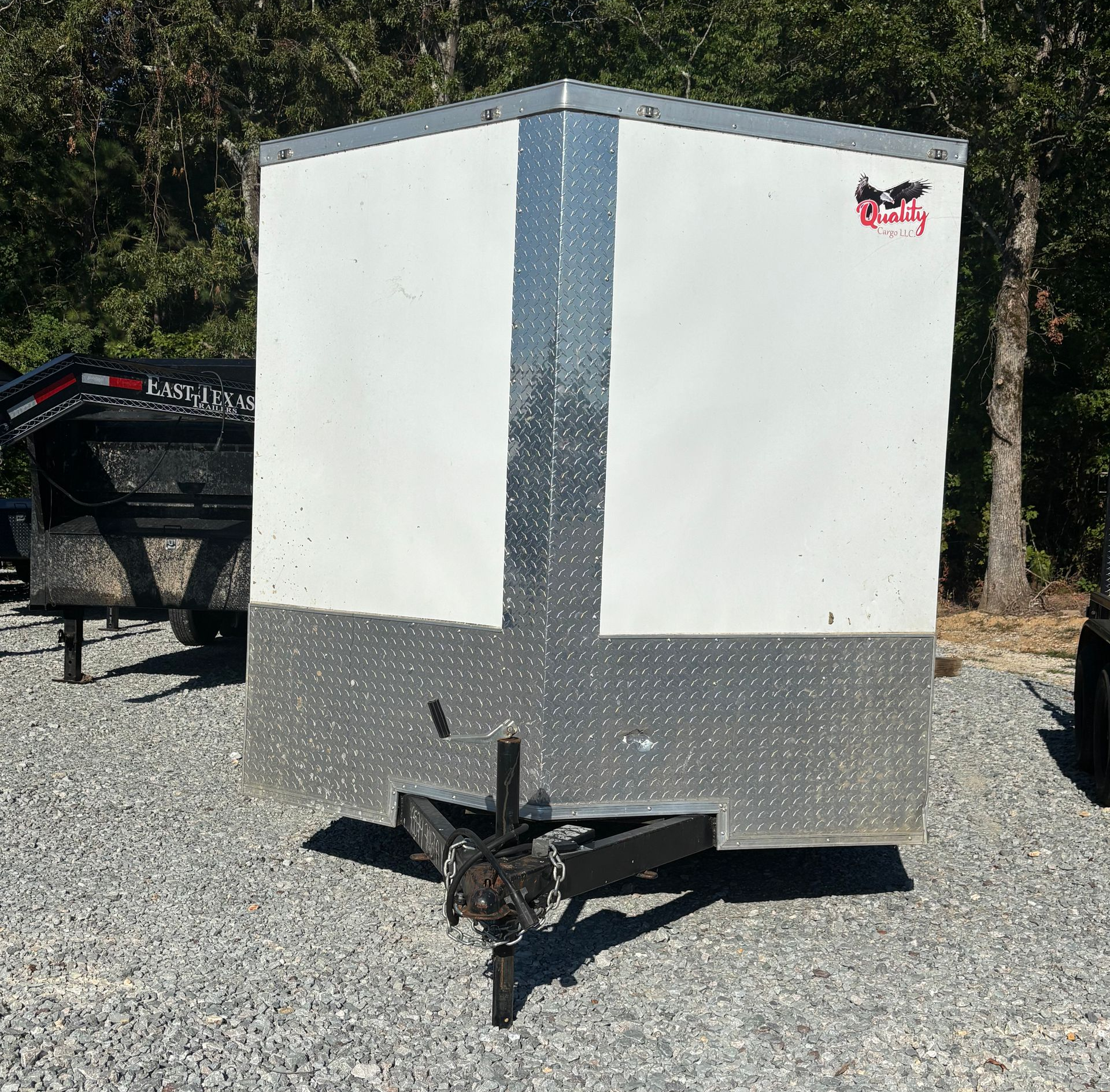 White enclosed cargo trailer on gravel with black hitch, surrounded by trees.