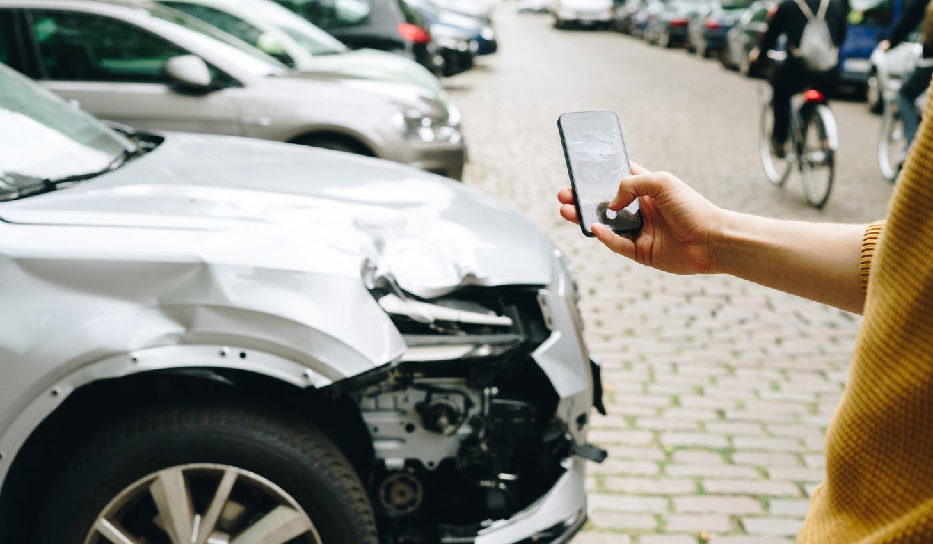 Person holding phone near a heavily damaged silver car on a cobblestone street.