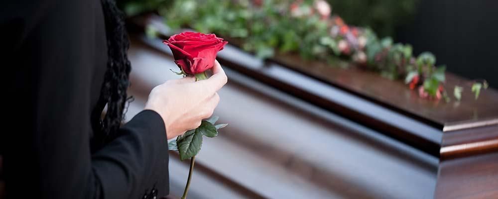 A woman is holding a red rose in front of a coffin.