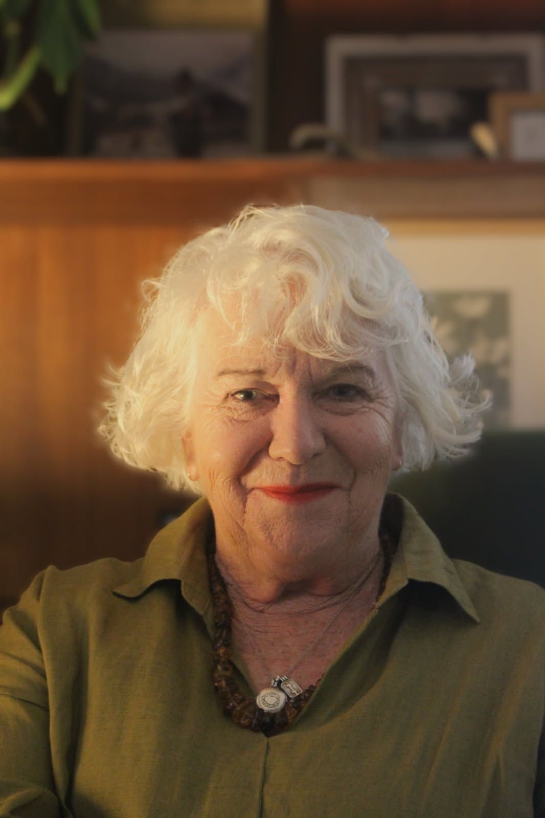 Smiling woman with curly white hair in an olive shirt, seated indoors with warm light and a blurred shelf behind her
