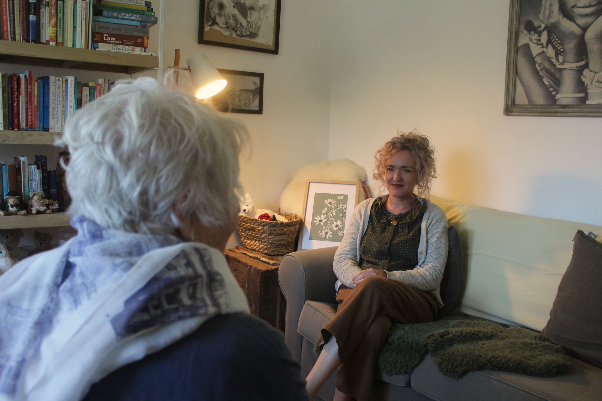 Two women talking in a cozy living room, with bookshelves, framed art, and soft lamp light.