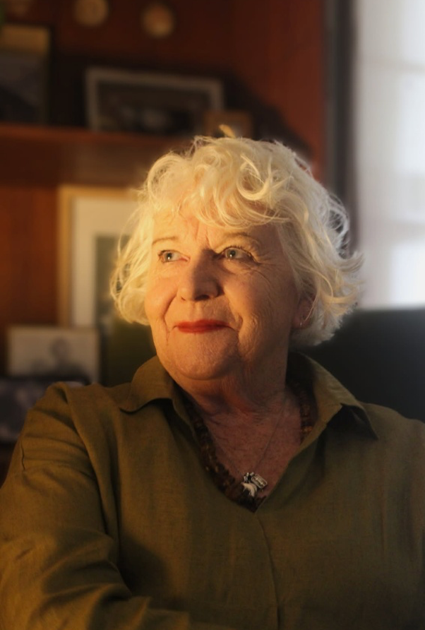 Portrait of a woman with curly white hair in warm indoor light, looking off to the side.