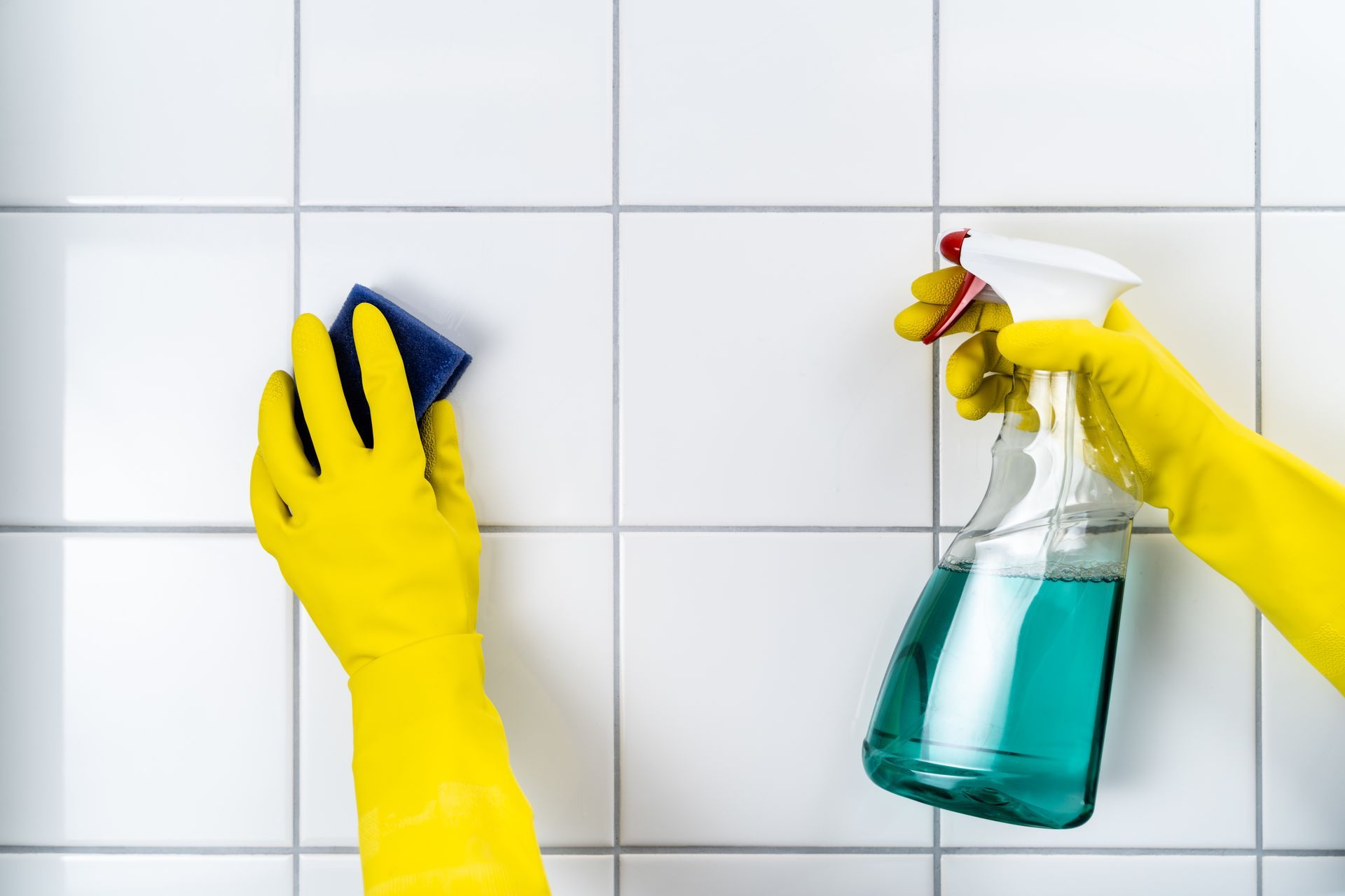 Hands in yellow rubber gloves scrubbing white tiled walls with a sponge and spray cleaner.