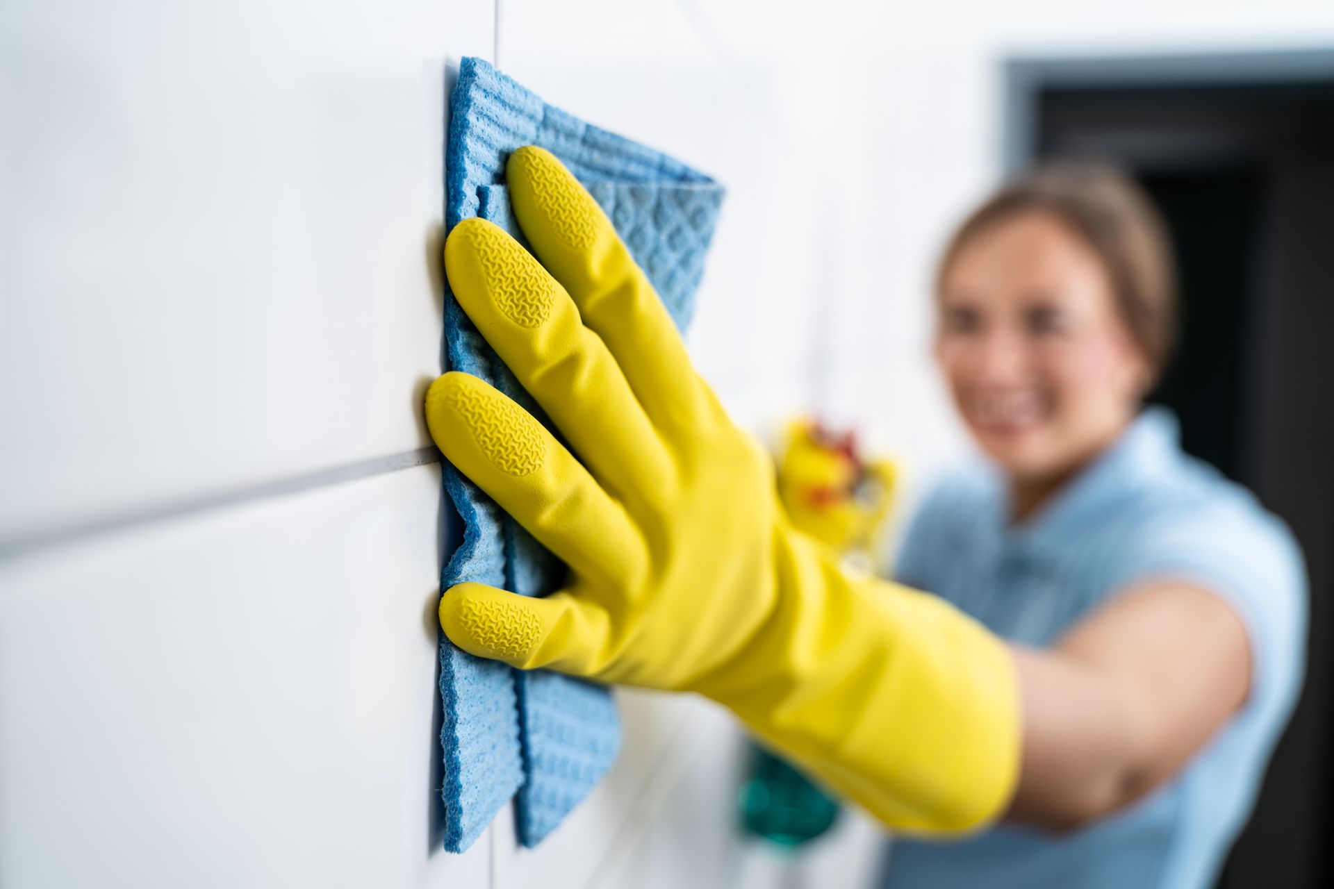 A hand in a yellow rubber glove holds a blue sponge, cleaning white tiles, with a person blurred in the background.