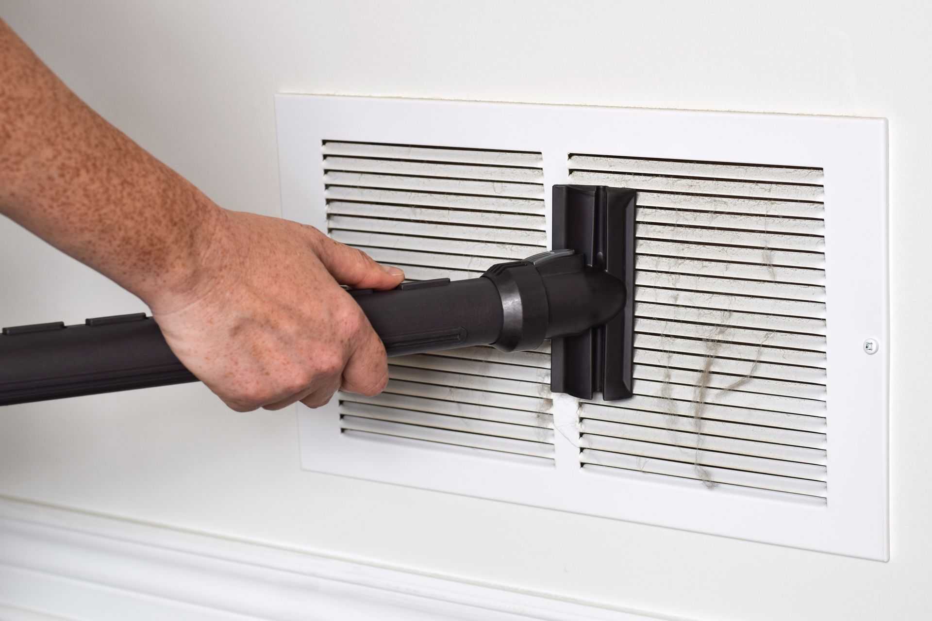 A hand uses a vacuum attachment to clean dust and debris from a white wall-mounted air vent grille.