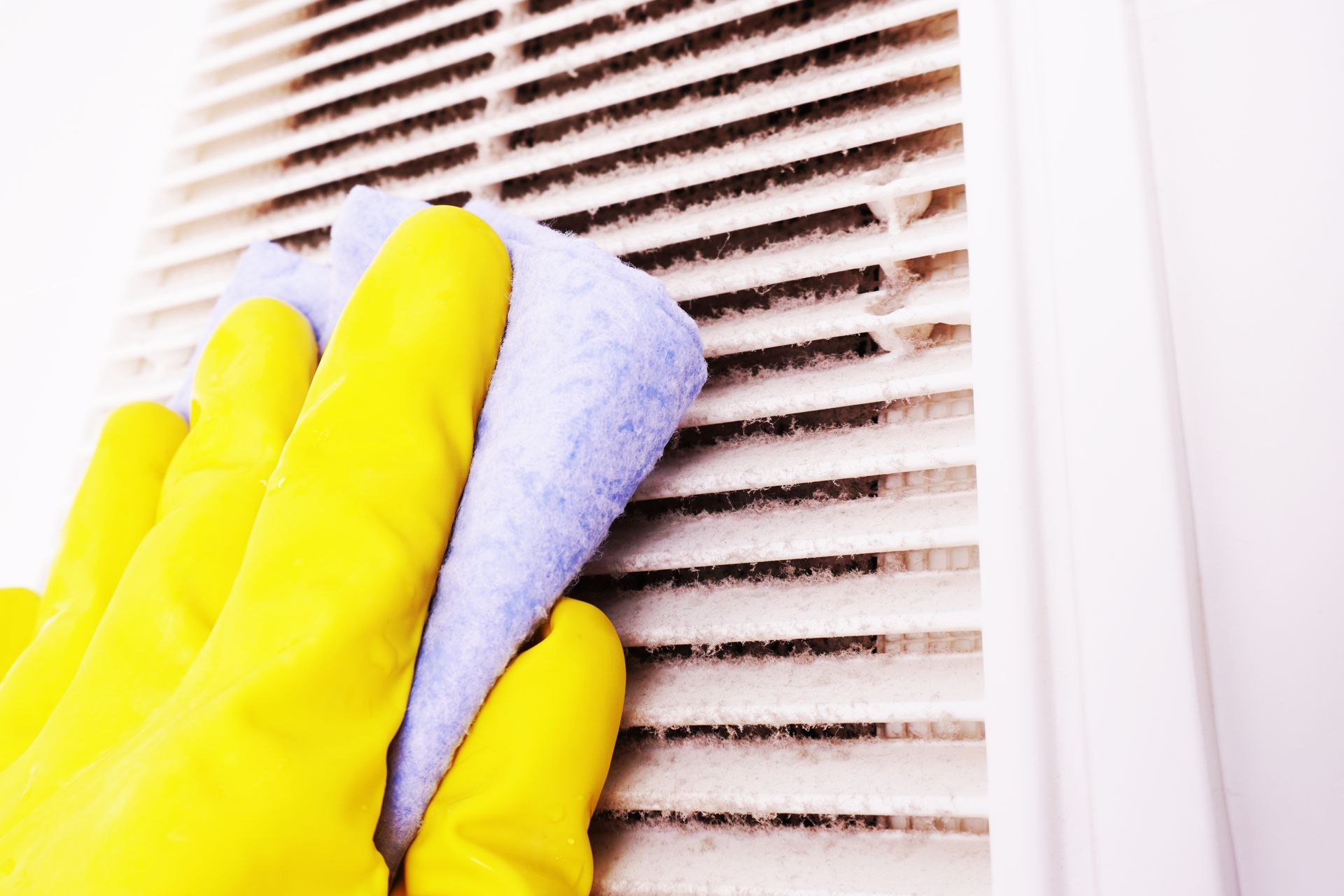 A hand wearing a yellow rubber glove uses a light blue sponge to clean dust from the slats of a white air vent.