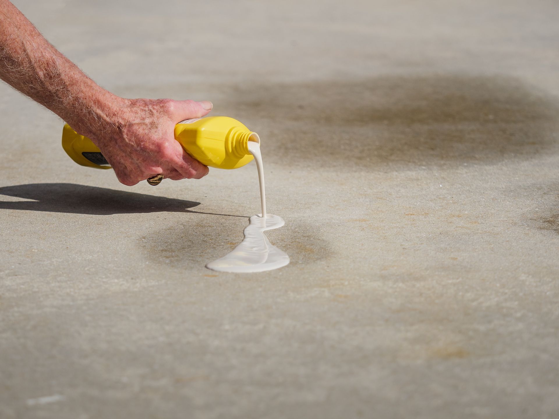 A person’s hand pours white liquid from a yellow bottle onto a gray concrete surface.