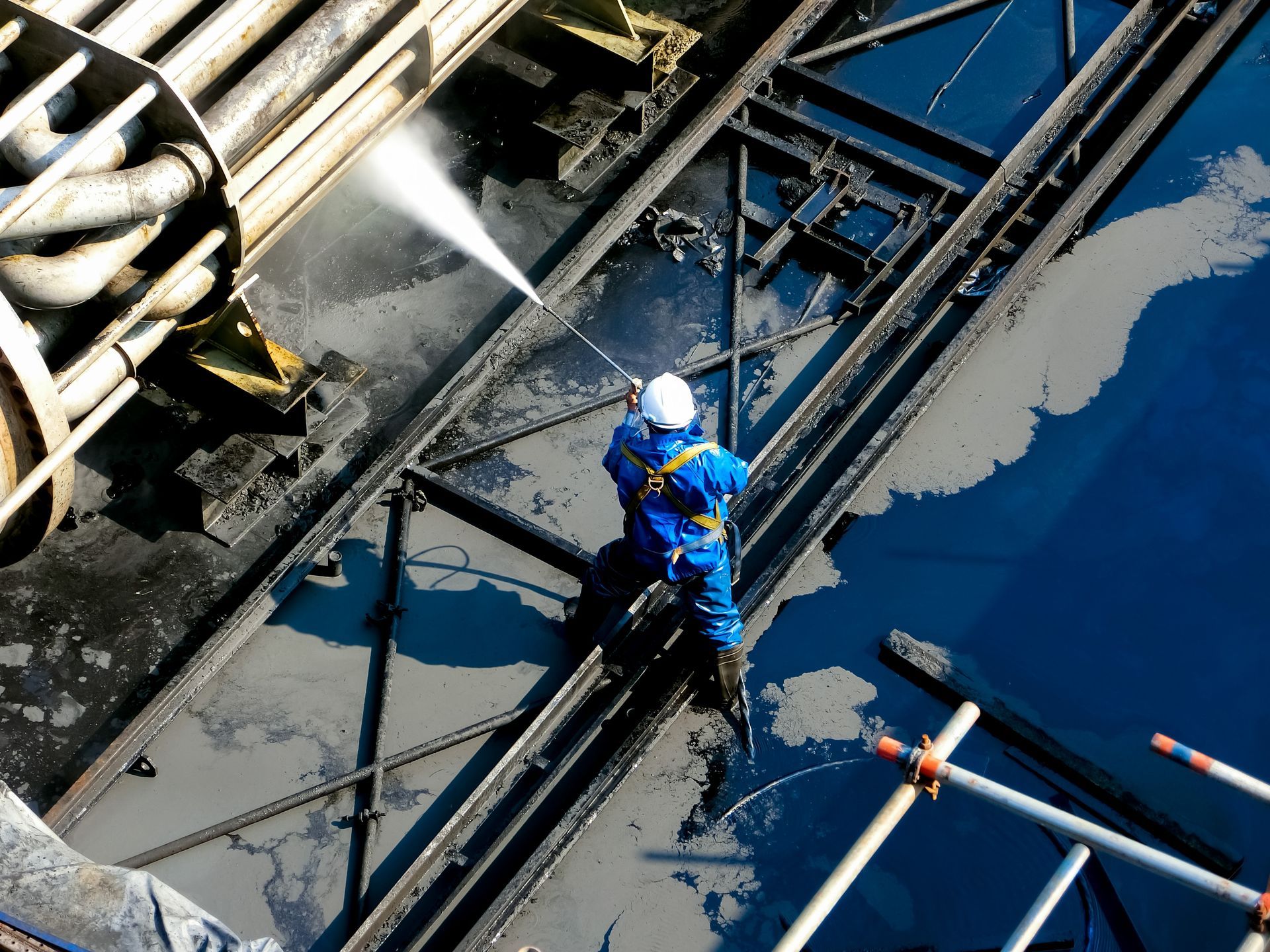 A worker in blue protective gear and a white hard hat uses a high-pressure hose to clean industrial metal structures.