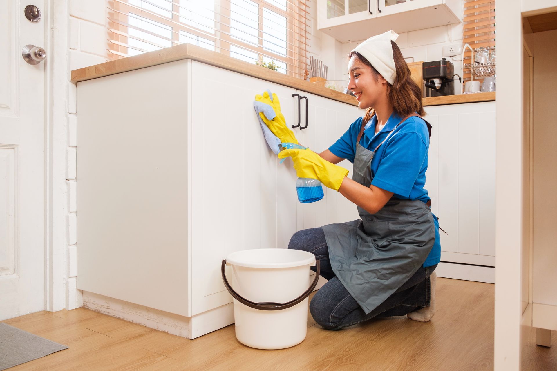 A professional cleaner wearing a blue uniform, apron, and yellow gloves wipes a white kitchen cabinet beside a bucket.