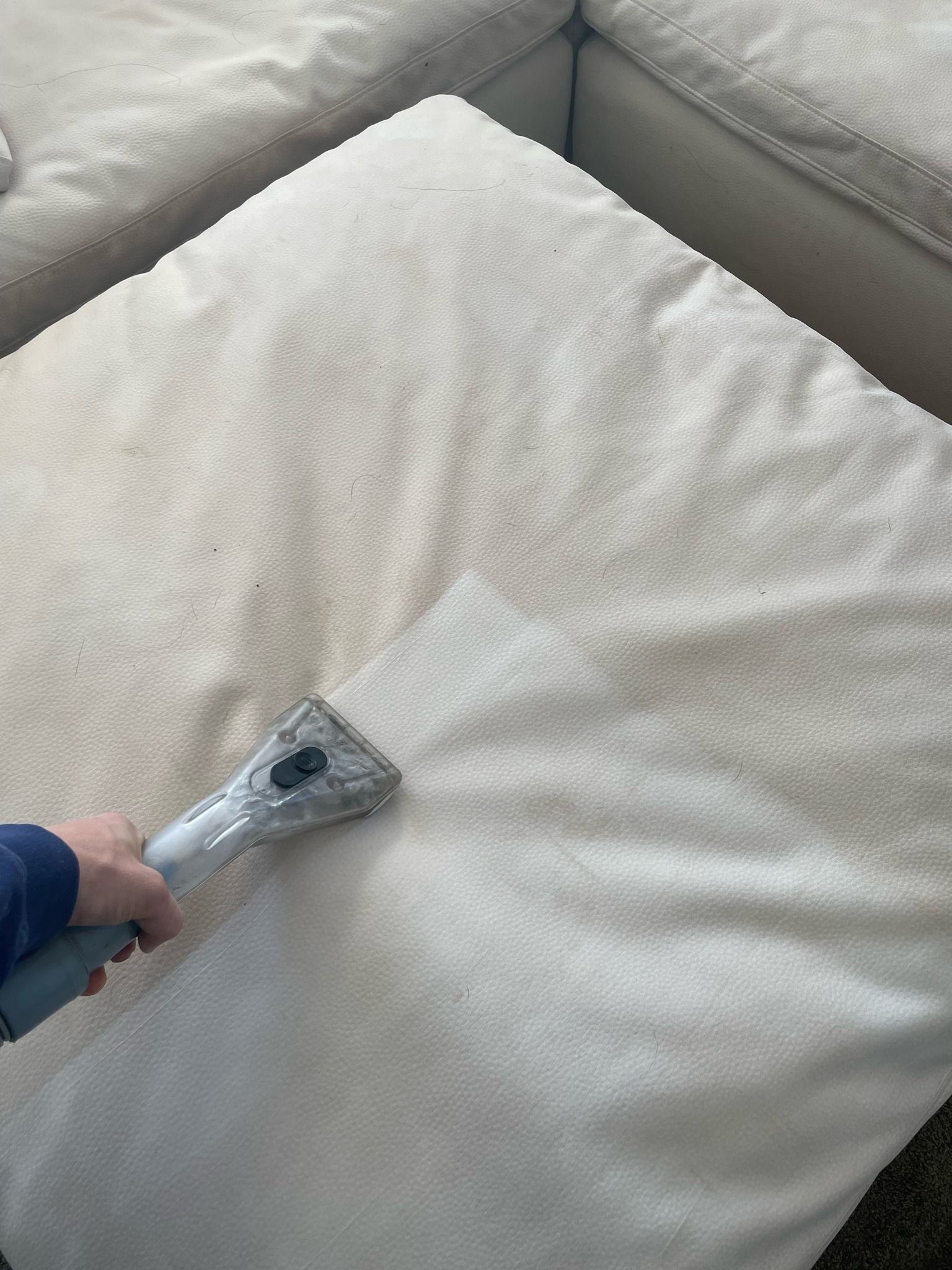 A person uses a handheld upholstery cleaner to deep clean a light-colored, textured fabric ottoman.