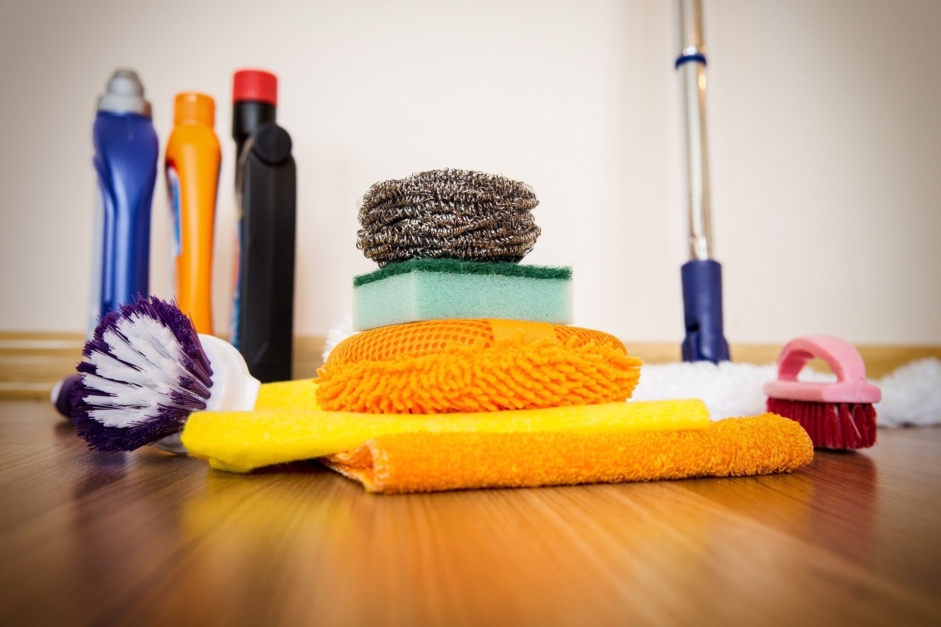 Cleaning supplies, including colorful bottles, sponges, brushes, and cloths, are stacked on a wooden floor.