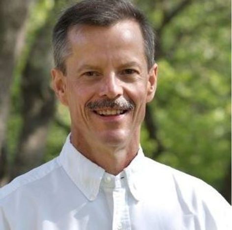 A man with a mustache is wearing a white shirt and smiling.