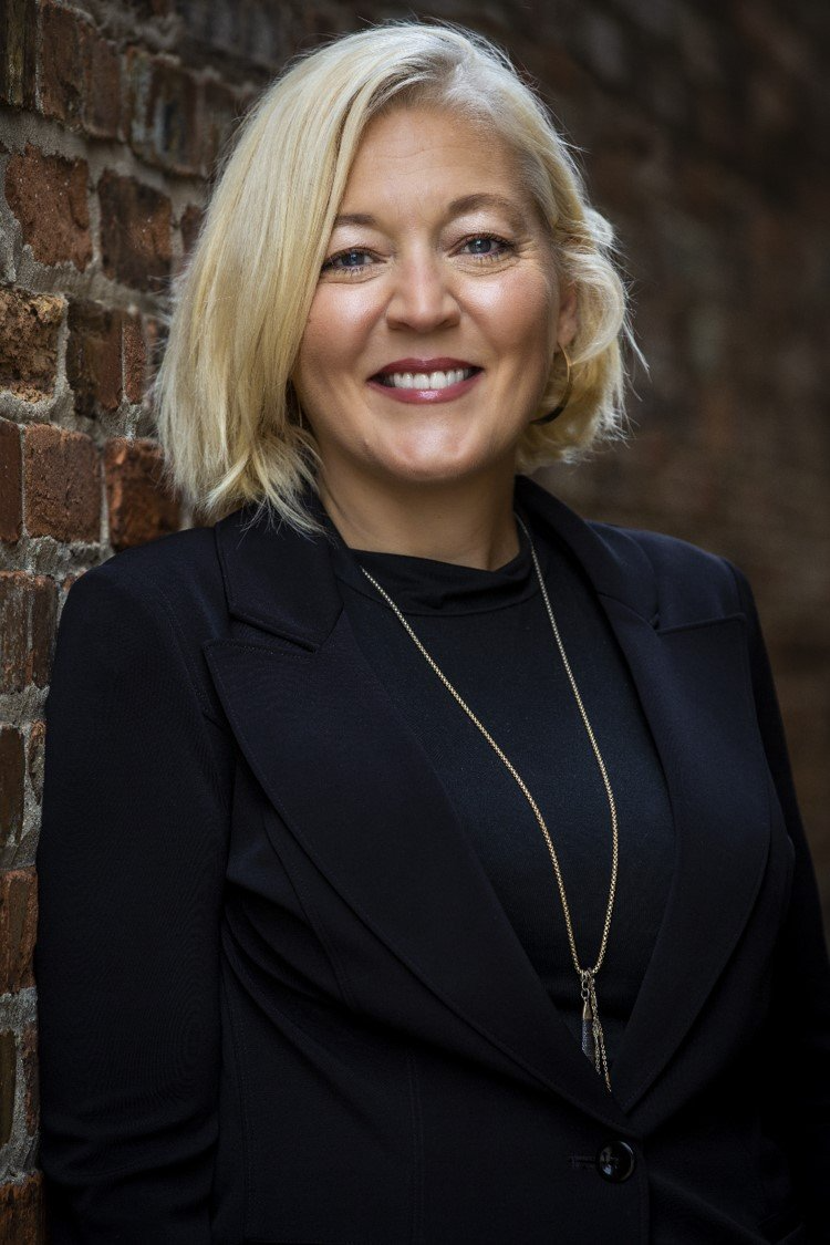 A woman in a black jacket and necklace is standing in front of a brick wall.