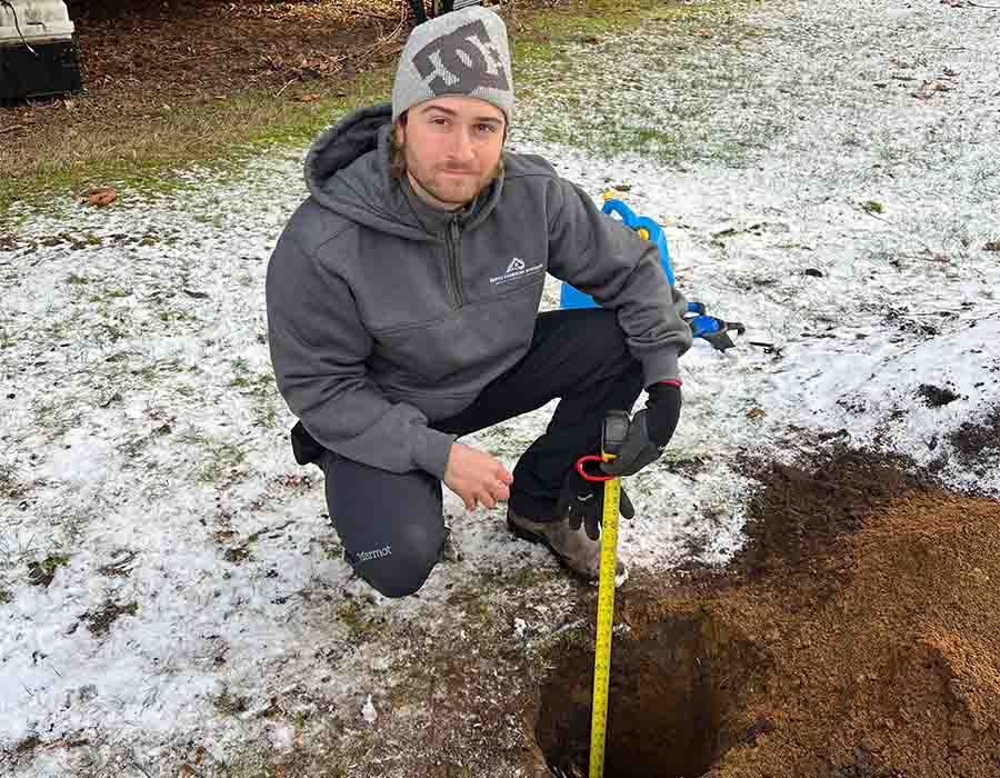 Person kneeling outdoors beside a yellow measuring tape, marking a snow-dusted patch of ground.