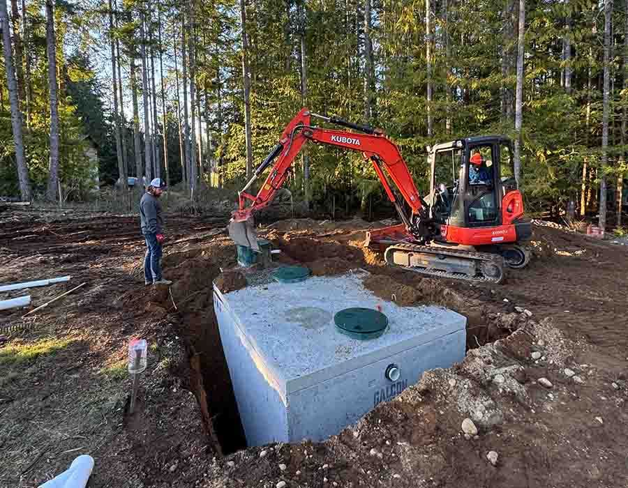 Red excavator installing a concrete tank at a wooded construction site, with a worker nearby.
