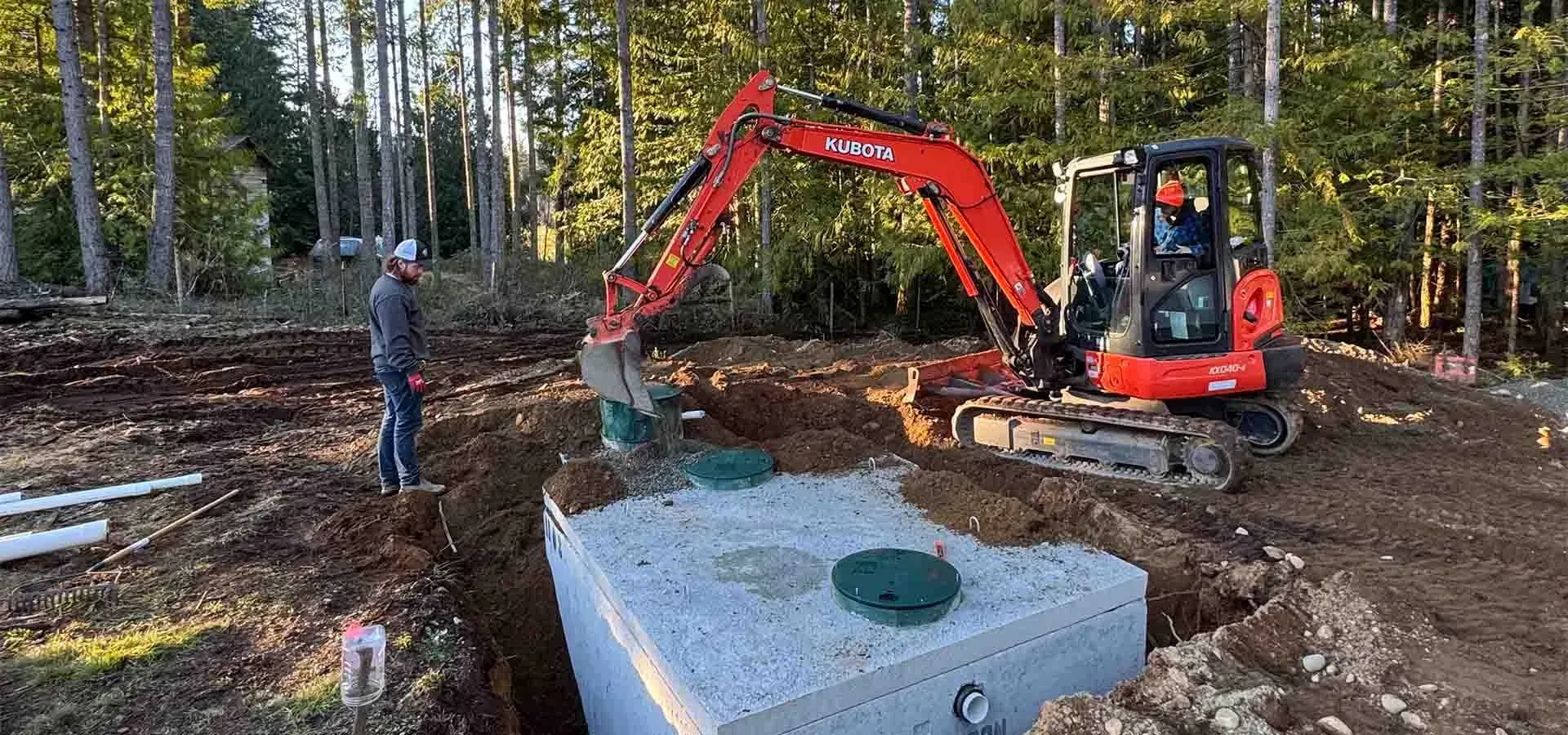 Red excavator lifting a concrete utility box on a muddy construction site with a worker nearby.