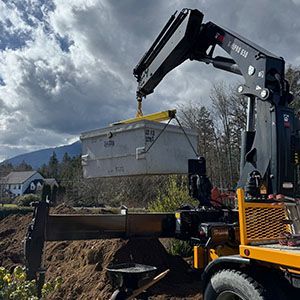 Crane lifting a gray metal container over a dump truck outdoors under cloudy skies