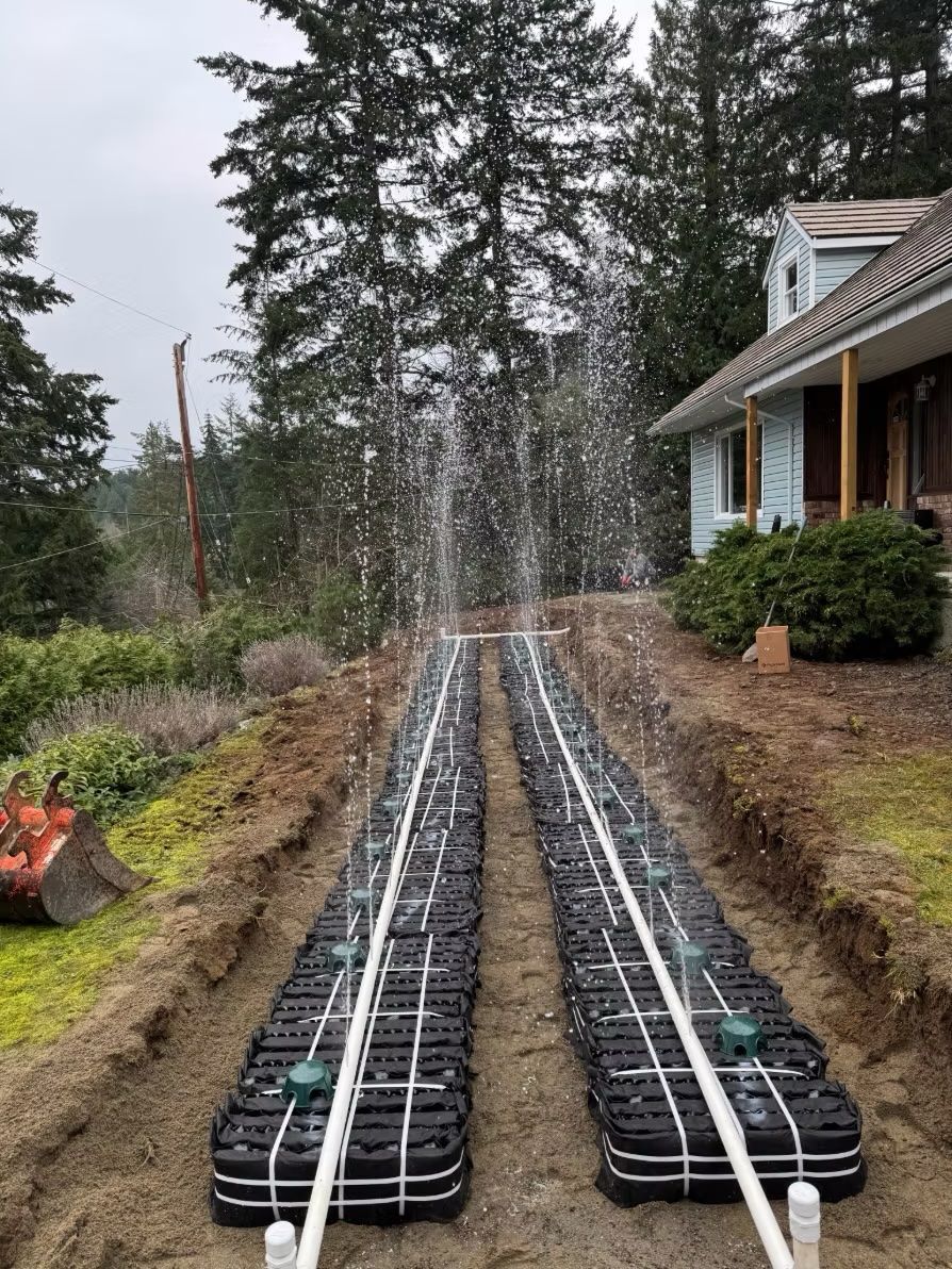Two black parallel trench drains spraying water beside a house in a wooded yard