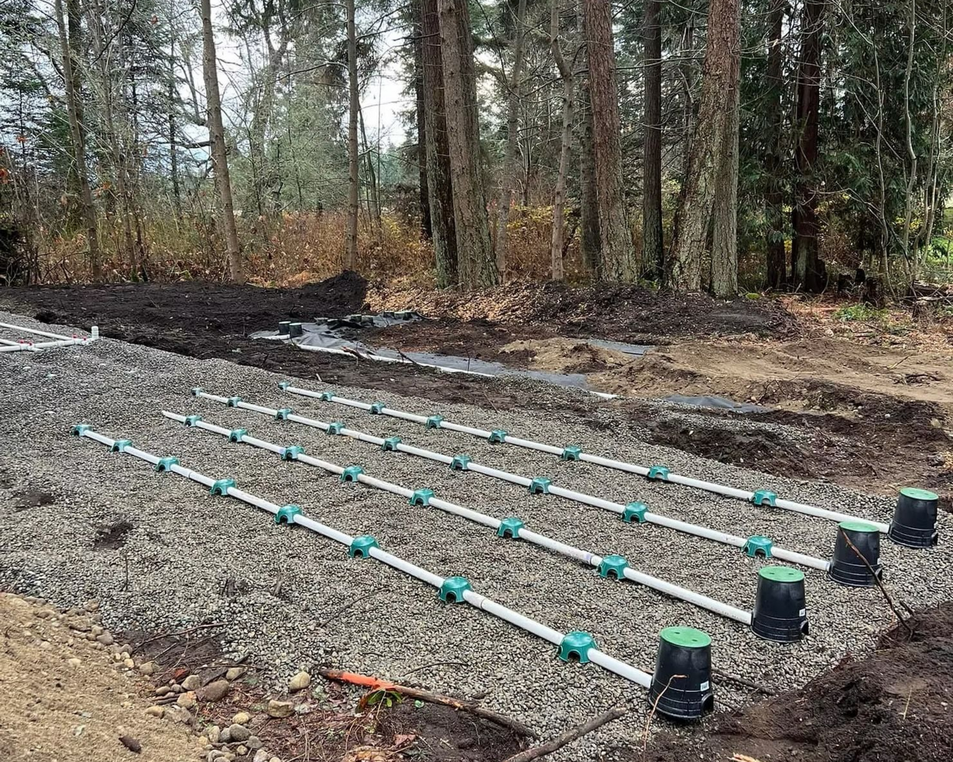 Raised turquoise pipes laid in parallel on gravel in a wooded clearing, with black buckets at one end