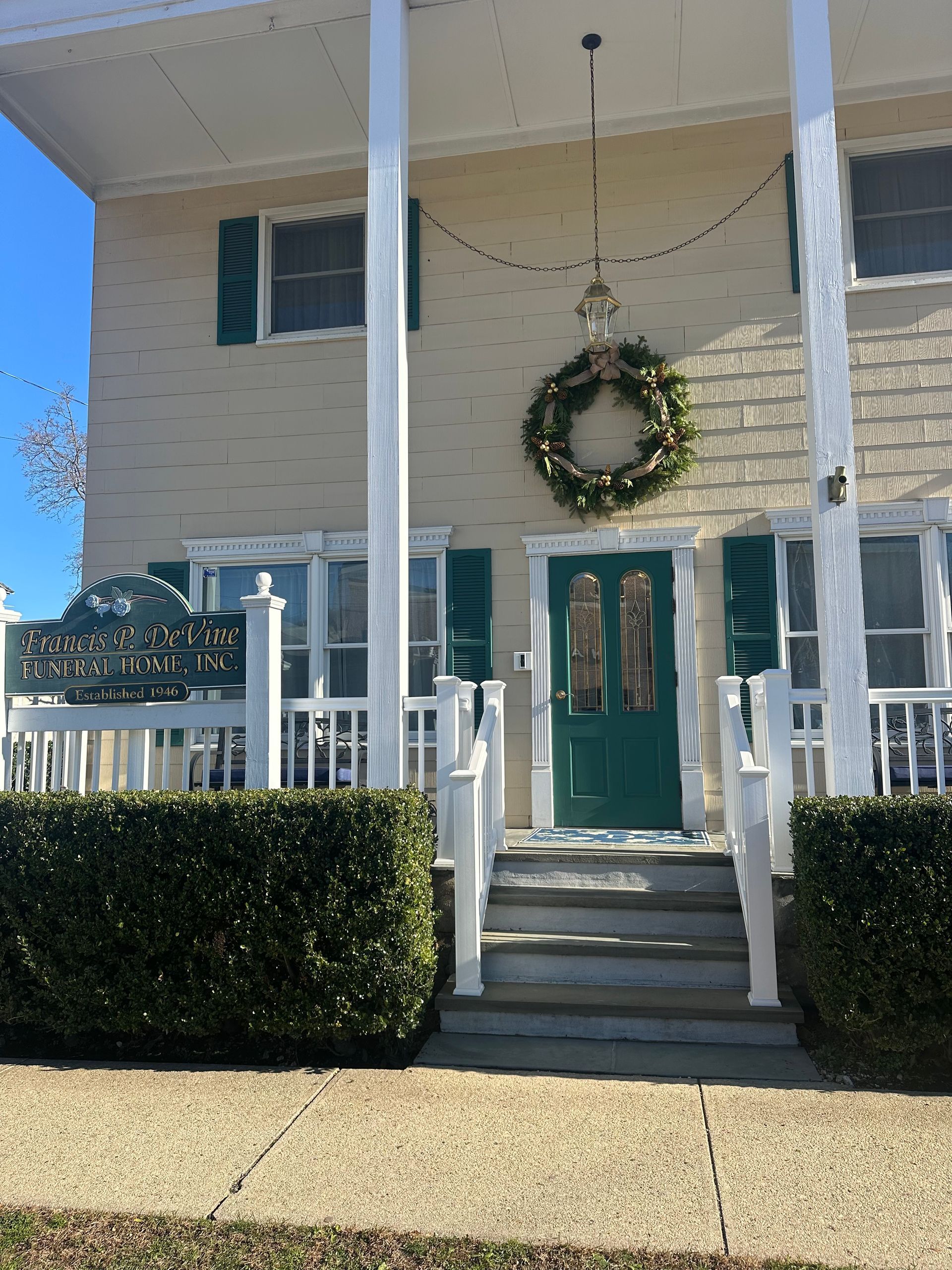 Two-story building with green door, shutters, and white porch. A wreath hangs on the door.