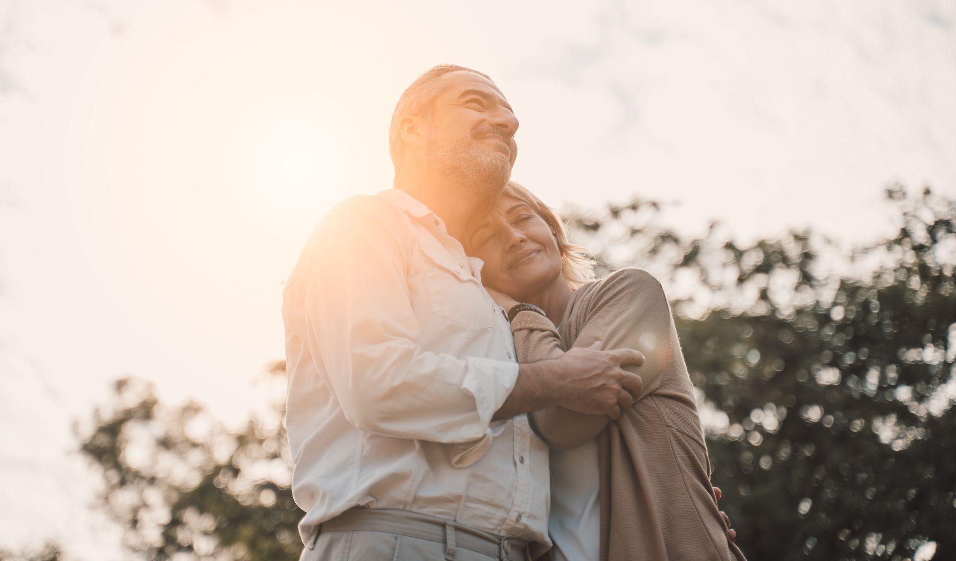 Elderly couple embracing outdoors, looking up. Sunlight shines on them.