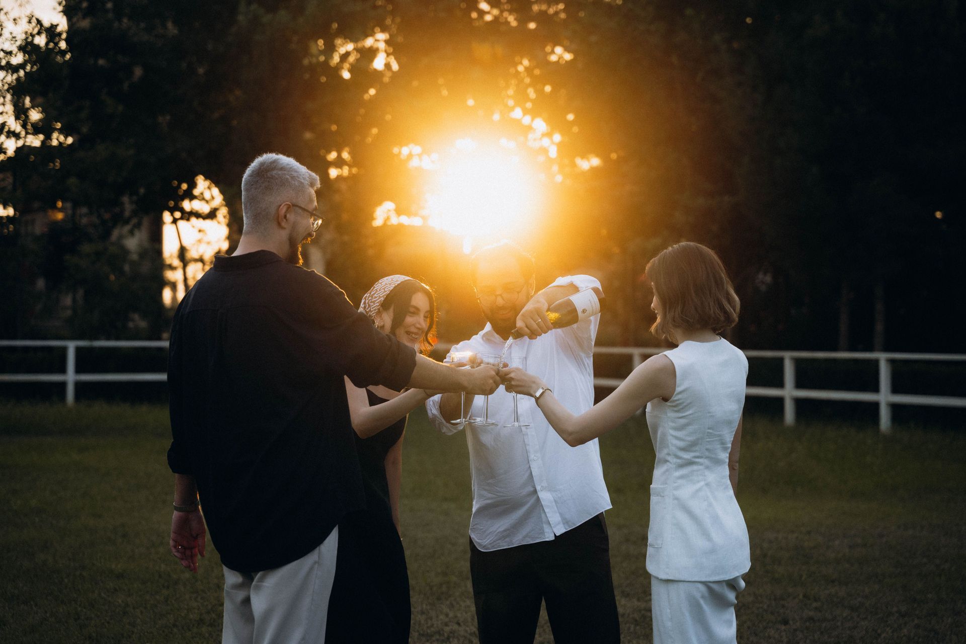 Four people holding hands in a circle at sunset in a park, with a bright sun flare behind them