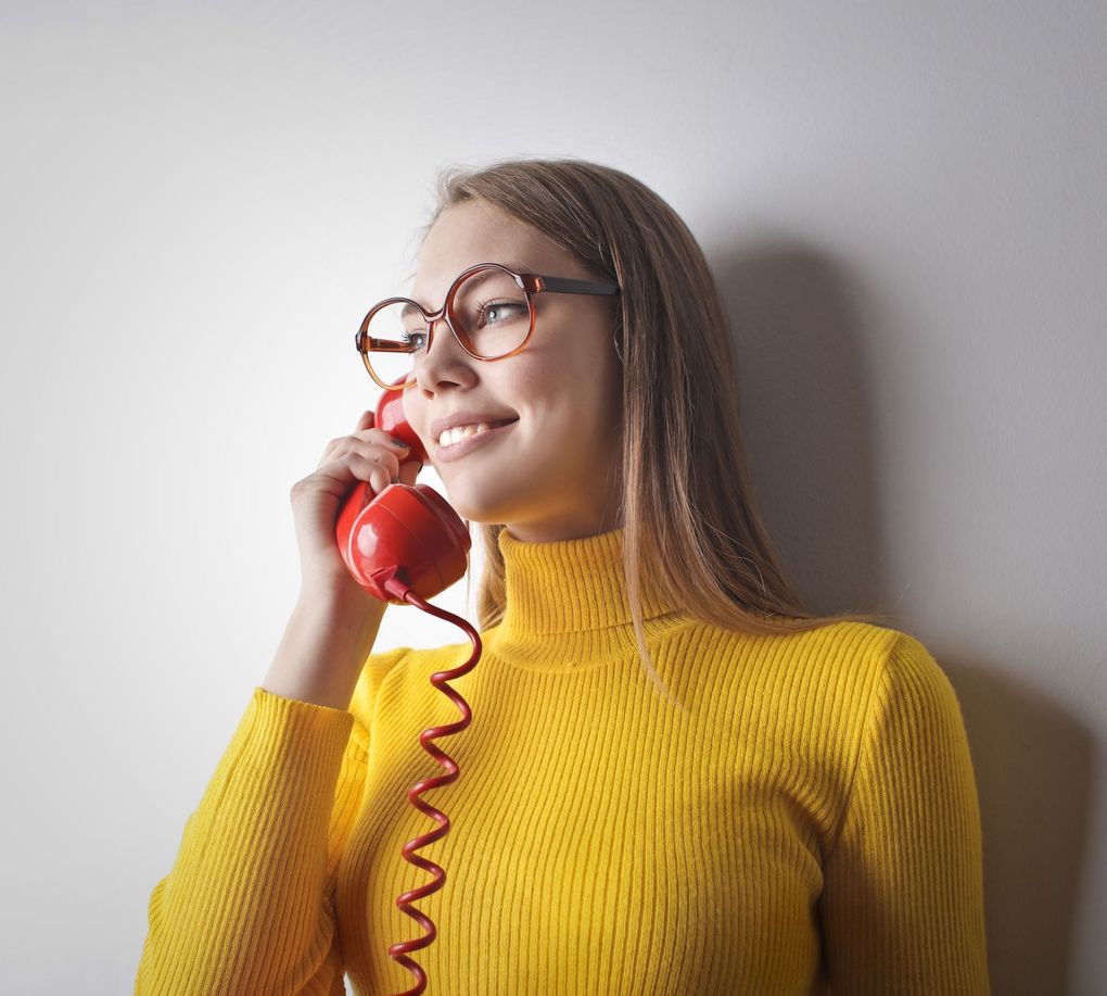 Smiling person in a yellow sweater holding a red corded phone against a gray wall
