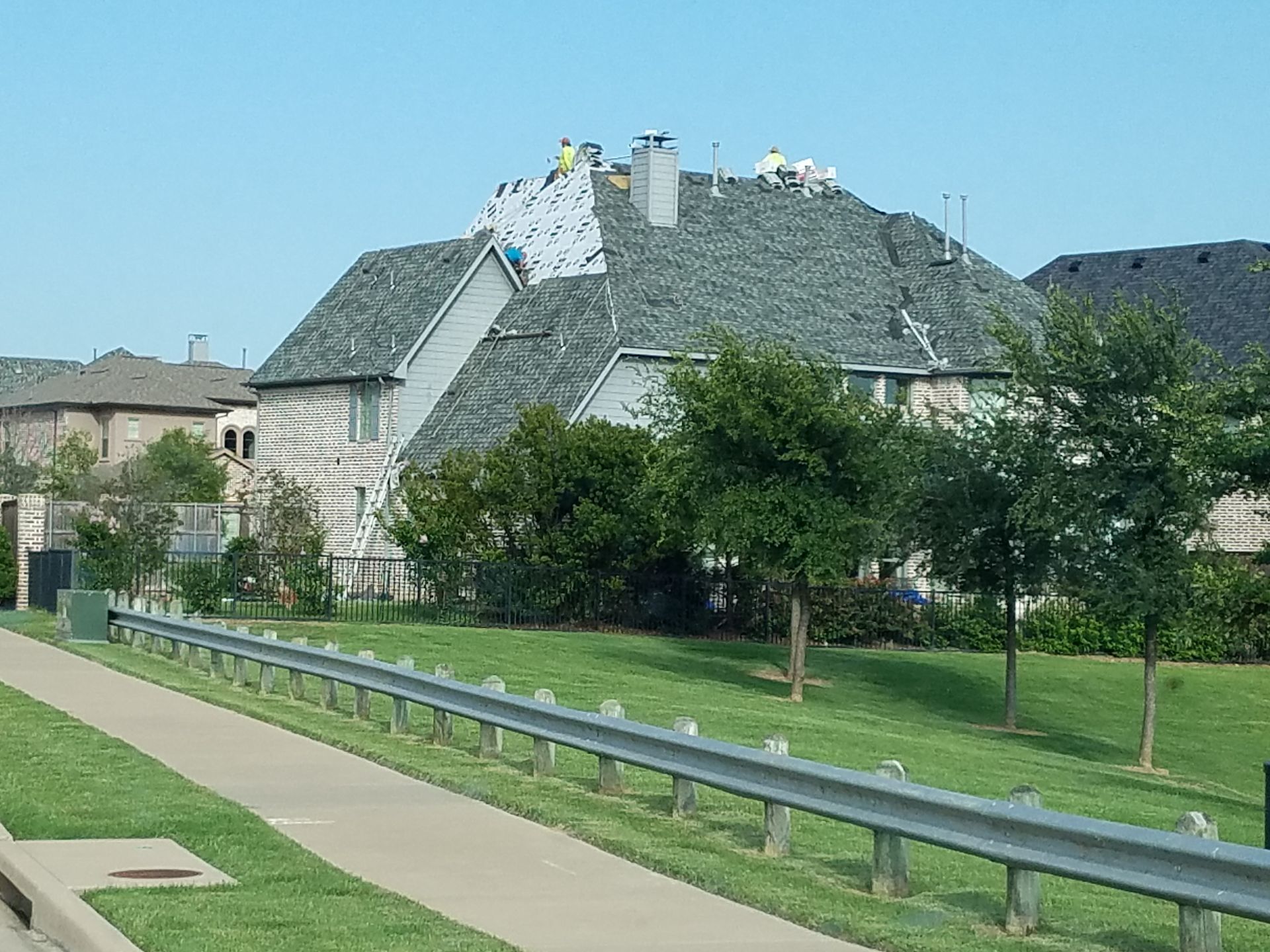 Roofers replacing shingles on a two-story house with a gray roof, blue sky, and green trees in front.