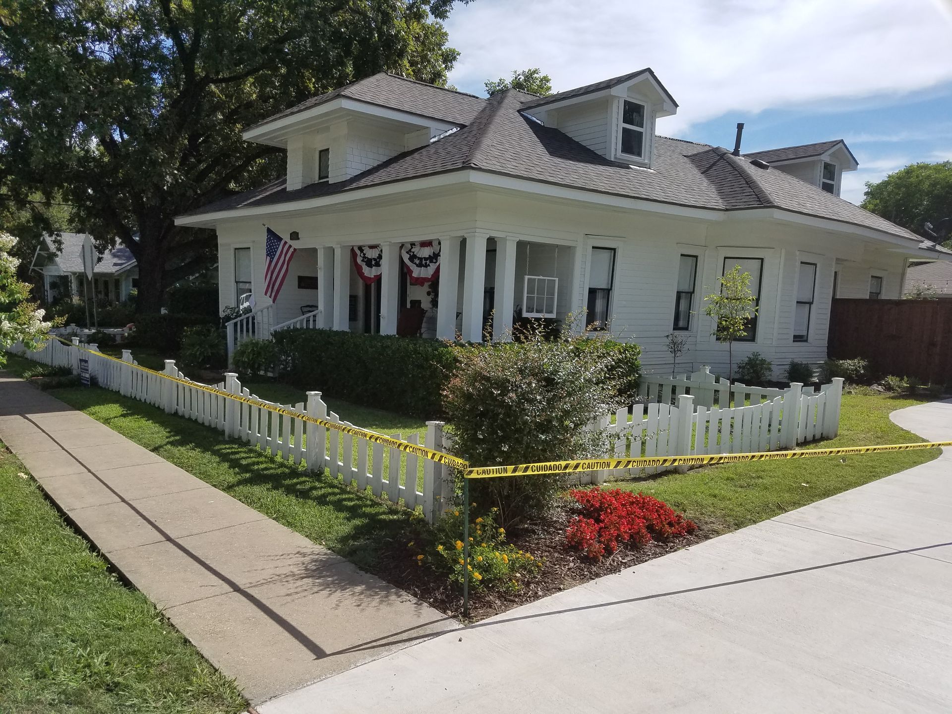 White house with picket fence, American flag, landscaping. Yellow caution tape marks off the yard.